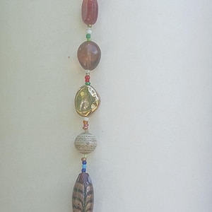 May include: A decorative wind chime featuring a variety of beads in different shapes and colours, suspended from a metal ring. The beads include gold, red, and purple, with a burgundy tassel at the base. The chime hangs against a white backdrop.
