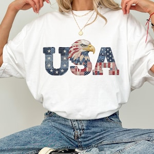 Puede incluir: Camiseta blanca con la palabra "USA" en un diseño patriótico. Las letras están decoradas con estrellas, rayas y un águila, en rojo, blanco y azul. La camiseta la lleva una persona con vaqueros azules.