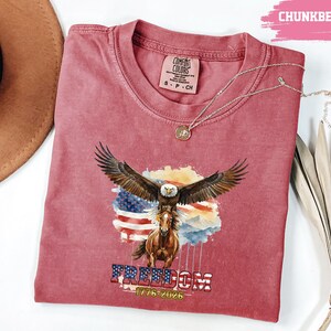 May include: A red Comfort Colors t-shirt with a graphic of an eagle with outstretched wings atop a horse, set against an American flag. The word "FREEDOM" is printed below, along with the dates "1776-2026". A silver necklace is draped across the shirt.