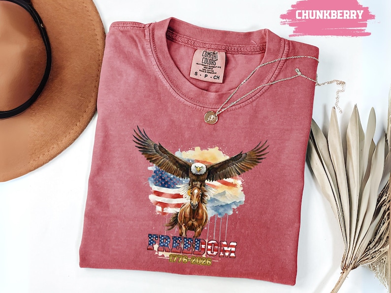 May include: A red Comfort Colors t-shirt with a graphic of an eagle with outstretched wings atop a horse, set against an American flag. The word "FREEDOM" is printed below, along with the dates "1776-2026". A silver necklace is draped across the shirt.