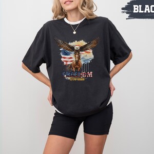 May include: Black t-shirt with a watercolour design of an eagle, horse, and American flag. The word "FREEDOM" is displayed below the image, along with the text "1776-2023".