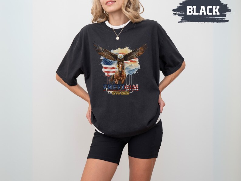 May include: Black t-shirt with a watercolour design of an eagle, horse, and American flag. The word "FREEDOM" is displayed below the image, along with the text "1776-2023".
