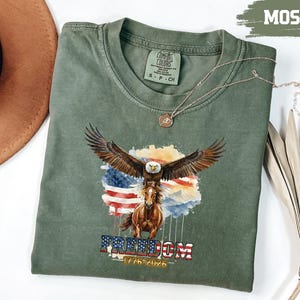 May include: A moss green t-shirt with a graphic of an eagle with outstretched wings above a horse, set against an American flag. The word "FREEDOM" and the dates "1776-2026" are below. A gold necklace is draped over the shirt. The shirt is a Comfort Colors brand.