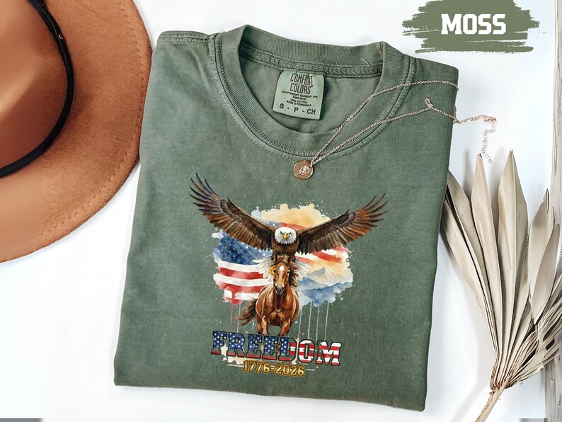 May include: A moss green t-shirt with a graphic of an eagle with outstretched wings above a horse, set against an American flag. The word "FREEDOM" and the dates "1776-2026" are below. A gold necklace is draped over the shirt. The shirt is a Comfort Colors brand.