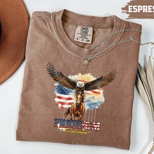 May include: Espresso-coloured t-shirt with a graphic of an eagle with outstretched wings above a horse, set against the American flag. The word "FREEDOM" and the dates "1776-2026" are below. A silver necklace is draped on the shirt. The shirt is a Comfort Colors brand.