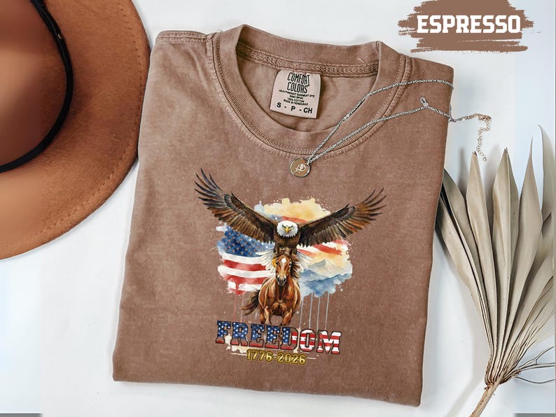 May include: Espresso-coloured t-shirt with a graphic of an eagle with outstretched wings above a horse, set against the American flag. The word "FREEDOM" and the dates "1776-2026" are below. A silver necklace is draped on the shirt. The shirt is a Comfort Colors brand.
