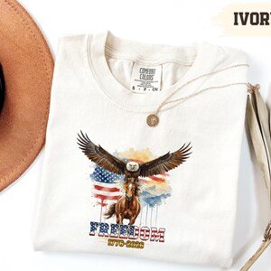 May include: Ivory-coloured t-shirt with a watercolour design of an eagle with outstretched wings above a horse, set against an American flag. The word "FREEDOM" is printed below, along with "1776-2026". A gold necklace is draped on the shirt.