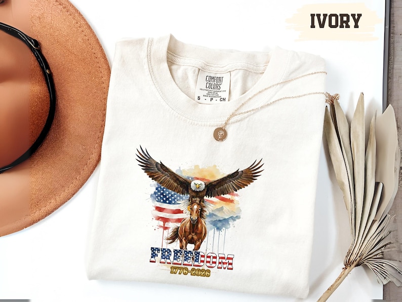 May include: Ivory-coloured t-shirt with a watercolour design of an eagle with outstretched wings above a horse, set against an American flag. The word "FREEDOM" is printed below, along with "1776-2026". A gold necklace is draped on the shirt.