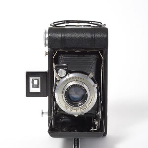 May include: A vintage black Kodak camera with a silver lens and a black leather handle. The camera has a rectangular viewfinder and a retractable bellows design. The lens has speed settings. The word "KODAK" is printed on the bottom.
