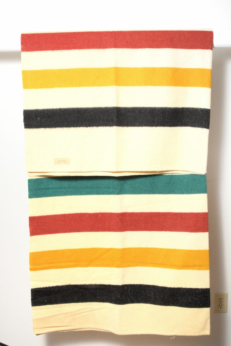 May include: A cream-colored blanket with horizontal stripes in red, yellow, black, and teal. The blanket is folded over a white bar. A small tag is visible on the blanket.