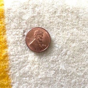 May include: A copper penny coin on a textured, off-white surface with a yellow stripe. The penny features a profile of Abraham Lincoln and the year 2010.