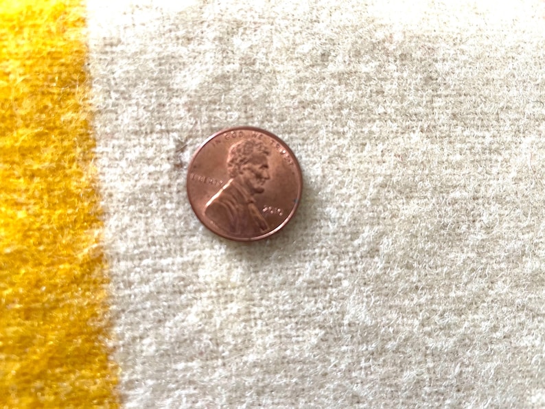 May include: A copper penny coin on a textured, off-white surface with a yellow stripe. The penny features a profile of Abraham Lincoln and the year 2010.