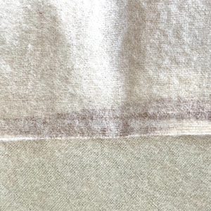 May include: Close-up of a textured, off-white fabric with a subtle, horizontal stripe in a muted brown hue. The fabric appears soft and possibly woven, with a slightly fuzzy texture. The image highlights the material's natural appearance.
