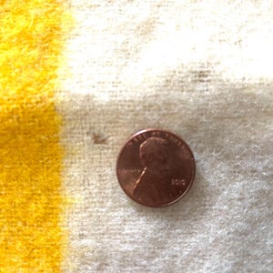 May include: A close-up of a penny on a yellow and white striped fabric. The penny is copper-colored and shows the profile of a person. The fabric has a textured, woven appearance.