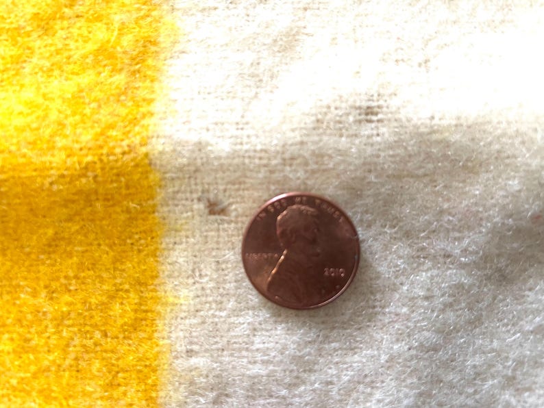May include: A close-up of a penny on a yellow and white striped fabric. The penny is copper-colored and shows the profile of a person. The fabric has a textured, woven appearance.