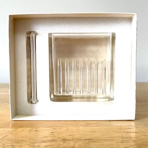 May include: A clear glass soap dish and a matching bar, presented in a white cardboard box. The soap dish has vertical ridges, and the bar is rectangular. The set is designed for bathroom use.