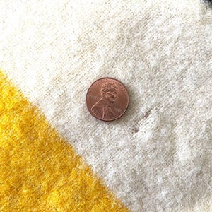 May include: Close-up of a textured, woven fabric with diagonal stripes in yellow, white, and black. A copper penny is placed on the fabric for scale. The fabric appears to be a blanket or throw.