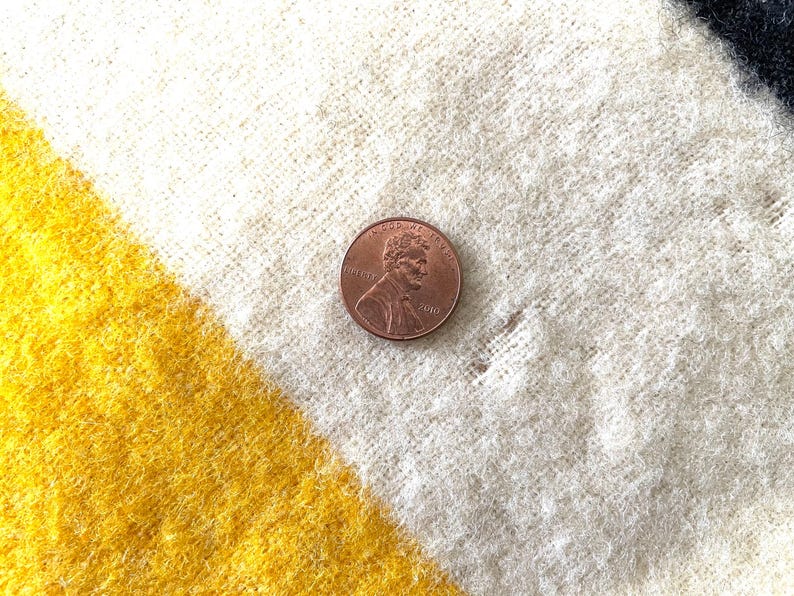 May include: Close-up of a textured, woven fabric with diagonal stripes in yellow, white, and black. A copper penny is placed on the fabric for scale. The fabric appears to be a blanket or throw.