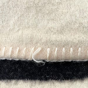 May include: Close-up of a textured, off-white fabric with a visible stitch detail along the edge. Below, a dark navy blue and black striped pattern is visible. The stitching is done with white thread.