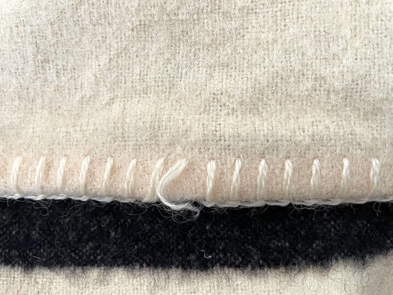 May include: Close-up of a textured, off-white fabric with a visible stitch detail along the edge. Below, a dark navy blue and black striped pattern is visible. The stitching is done with white thread.