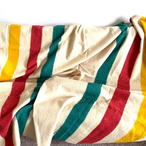 May include: A cream-colored blanket with vertical stripes in black, yellow, red, and teal. The blanket is folded and draped, showcasing the colorful stripes. This cozy blanket is perfect for adding warmth and style to any space.