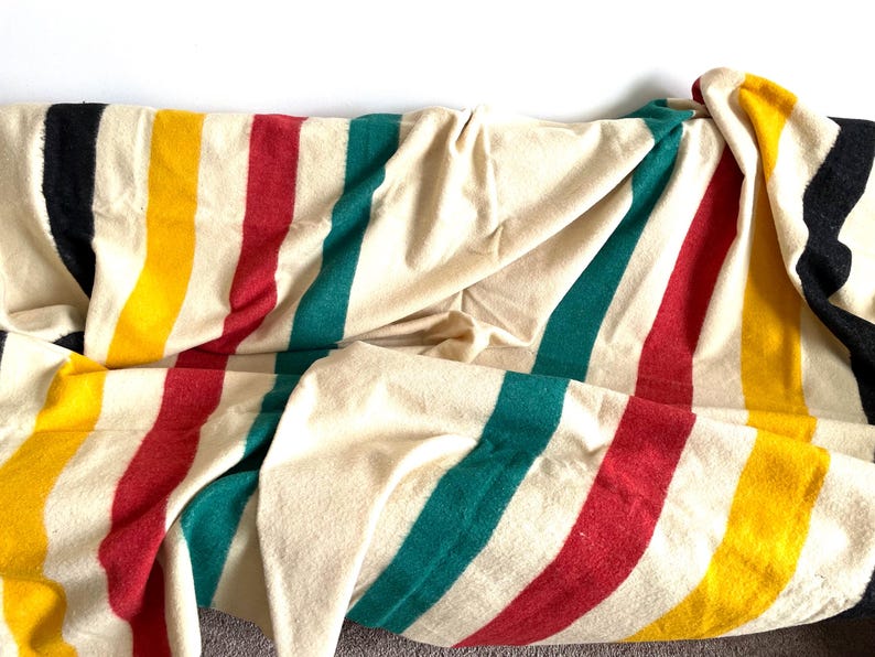 May include: A cream-colored blanket with vertical stripes in black, yellow, red, and teal. The blanket is folded and draped, showcasing the colorful stripes. This cozy blanket is perfect for adding warmth and style to any space.