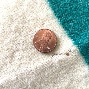 May include: A copper penny rests on a textured fabric with white, teal, and off-white stripes. The penny shows the profile of a man and the words "In God We Trust" and the year 2010.