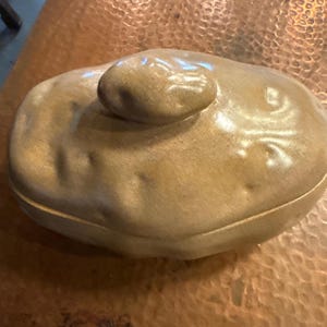 May include: A tan ceramic lidded dish with a rounded, potato-like shape. The lid features a small, rounded knob. The dish has a textured surface with subtle variations in color, suggesting a handmade quality. The overall design is organic and rustic.