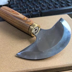 K&ouml;nnte beinhalten: Ein gebogenes Lederwerkzeugmesser mit Holzgriff und Messingzwinge. Die Klinge besteht aus poliertem Stahl. Der Griff hat ein Holzmaserungsmuster.