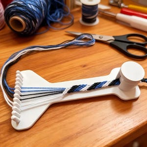 Puede incluir: Una herramienta de tejido de plástico blanco con un poste cilíndrico en un extremo y múltiples clavijas en el otro. Un cordón parcialmente trenzado en negro, blanco y tonos de azul está sujeto al telar, con hilos sueltos en las clavijas.