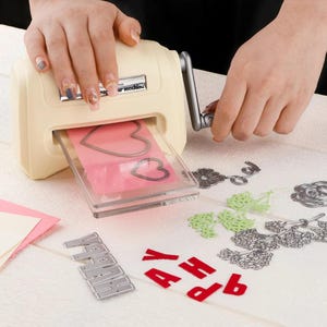 May include: A cream-coloured manual die-cutting machine with a grey crank handle. A clear plastic plate containing two metal heart-shaped dies and a pink paper sheet is inserted into the machine. Metal floral and letter dies are arranged nearby.