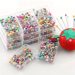 Könnte beinhalten: Eine Sammlung rechteckiger Aufbewahrungsboxen aus klarem Kunststoff, gefüllt mit Stecknadeln mit bunten, perlenartigen Köpfen. Ein rundes rotes Nadelkissen aus Stoff mit einem grünen, sternförmigen Filzaufsatz befindet sich rechts daneben.