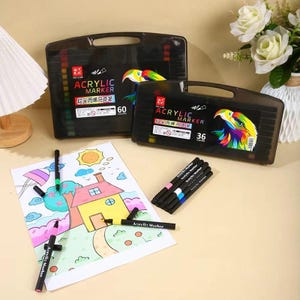 Può includere: Due custodie in plastica nera per pennarelli acrilici, una etichettata con 60 colori e l'altra con 36 colori. Diversi pennarelli neri singoli sono posizionati su un foglio di carta bianco con un disegno colorato di una casa e un sole.