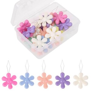 Puede incluir: Una caja de almacenamiento rectangular de plástico transparente llena de enhebradores de agujas, cada uno con un mango de plástico de colores en forma de flor. Cinco enhebradores individuales en rosa, azul, melocotón, morado y blanco están alineados.