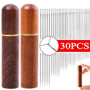 Può includere: Due astucci per aghi in legno, uno marrone scuro e uno marrone chiaro, con dettagli dorati, accanto a un set di aghi da cucito. Un banner rosso mostra "30PCS". Gli aghi argentati sono sottili, ideali per cucire e creare.