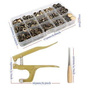 May include: A clear plastic storage box containing various bronze-colored metal snap fasteners, a gold-toned metal hand press tool with a spring mechanism, and a metal punch tool with a wooden handle, with dimensions labeled in mm and inches.