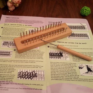 Peut inclure: Un métier à tricoter rectangulaire en bois avec deux rangées parallèles de broches en métal et un crochet assorti à manche en bois. Le métier et l'outil sont posés sur une feuille d'instructions en papier avec du texte et des schémas.