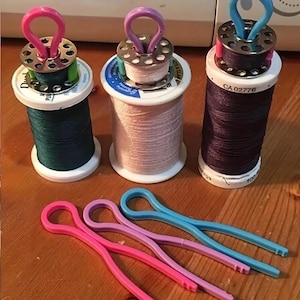May include: Three sewing thread spools with metal bobbins attached to the top, featuring pink, purple, and blue plastic bobbin buddies. Below are three matching plastic bobbin buddies in pink, purple, and blue, designed to hold thread ends.