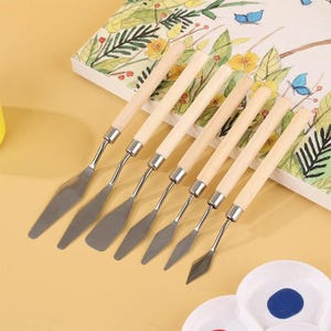 May include: A set of seven painting palette knives with light-colored wooden handles and silver-toned metal blades of varying shapes and sizes, arranged in a row on a flat surface next to a sketchbook with floral illustrations and a paint palette.