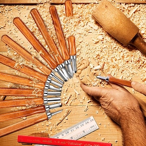 May include: A set of wood carving tools with polished wooden handles and metal blades, arranged in a fan pattern. A wooden mallet, a ruler with imperial markings, and a red pencil with text are included alongside a carved wooden piece.