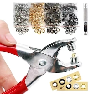 Puede incluir: Una herramienta de metal para colocar ojales con mangos rojos y mandíbulas plateadas, acompañada de cuatro bolsas de plástico con ojales metálicos plateados, dorados, bronce y negros, un punzón y dos tiras adhesivas con muestras de ojales.