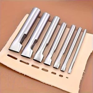 May include: A set of seven silver-colored metal hole punches arranged in descending order of size, positioned diagonally across a piece of light beige leather. Each tool features a cylindrical handle and a flat, hollow cutting end for creating slots.