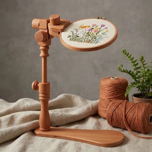 Puede incluir: Un soporte de madera de color marrón claro para bastidor de bordado con brazo y base ajustables. Un bastidor circular está sujeto en la parte superior, sosteniendo una tela color crema con un diseño floral bordado de flores silvestres.