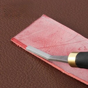 May include: A metal leathercraft creaser tool with a black handle and brass ferrule, positioned on a rectangular strip of red leather. The tool features a thin, angled metal tip resting against the edge of the textured red leather material.