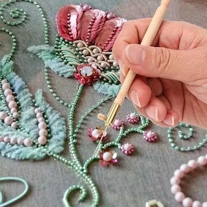 May include: A tambour embroidery hook with a light wood handle and a gold metal tip, used to stitch intricate floral patterns onto fabric. The design features pink sequins, light green beads, and small white pearls arranged in a botanical motif.