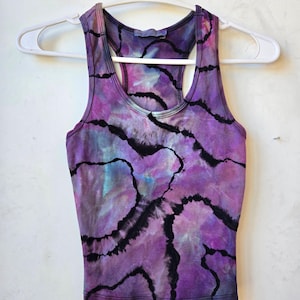 Pode incluir: Top sem mangas roxo tie-dye com detalhes lineares pretos. O top estilo nadador apresenta decote redondo e está pendurado em um cabide branco. O tecido tem uma mistura de tons roxos, azuis e rosa.