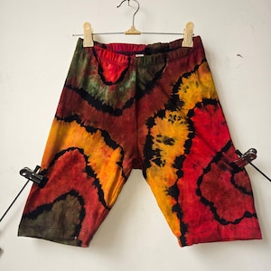 May include: Tie-dye shorts in a vibrant mix of red, orange, yellow, and green hues, with black accents. The shorts are hanging on a hanger against a white background. The fabric appears soft and comfortable.