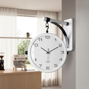 May include: A white, double-sided quartz clock with black hands and numerals. The clock is mounted on a white bracket with a black curved arm. The clock's diameter is approximately 10 inches.