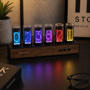 Puede incluir: Un reloj Gixie con seis tubos de vidrio verticales que muestran números iluminados en azul, morado, magenta, rojo y amarillo. La base es de madera con un acabado oscuro y cuenta con tres botones de control circulares en el lado derecho.