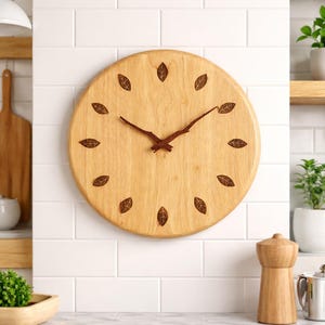 Op de afbeelding: Een ronde houten wandklok met een lichtbruine afwerking. De wijzerplaat heeft donkerbruine bladvormige markeringen en bijpassende wijzers. De klok is gemonteerd op een witte betegelde muur, wat wijst op een keuken of eetkamer.